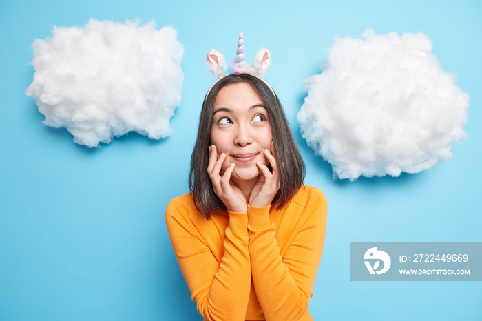 Portrait of pleased young Asian woman looks with dreamy expression keeps hands on face smiles gently recalls pleasant memories wears unicorn headband and orange jumper isolated over blue wall