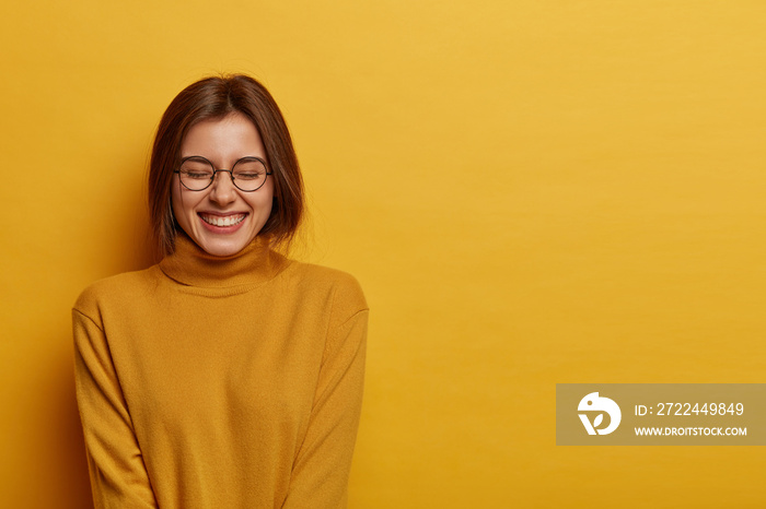 Feminine girl with toothy smile, squints face and feels glad, expresses positive emotions, wears casual long sleeved jumper, isolated on yellow background, laughs happily, gets pleasant words.