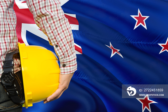 Engineer is holding yellow safety helmet with waving New Zealand flag background. Construction and building concept.