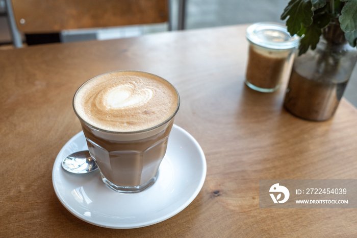 Flat white coffee in clear transparent glass and white plate and flower on the vase, on rough wooden table beside windows blur background of sidewalk.