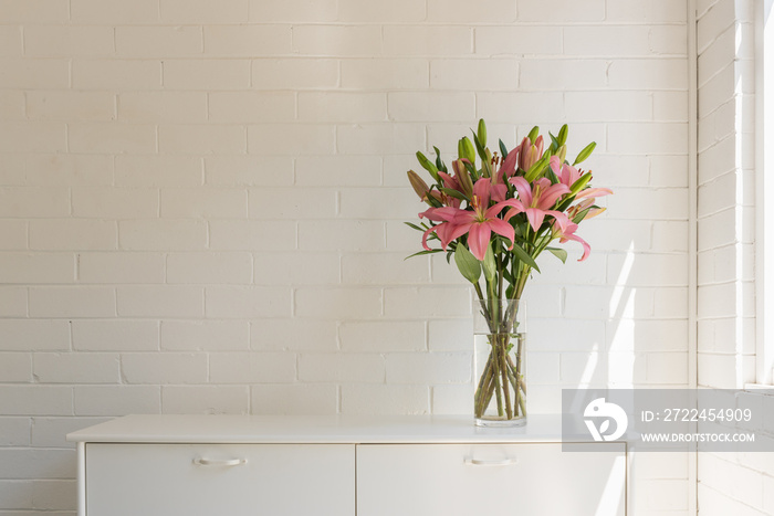 Pink asiatic lilies in glass vase on white sideboard against painted brick wall with sunlight (selective focus)