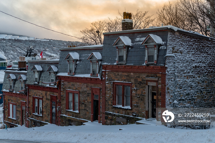 Multiple attached buildings on a hill with brown rock walls, double hung windows and chimneys. The vintage buildings have snow in front of them on the ground and the sun setting in the background.