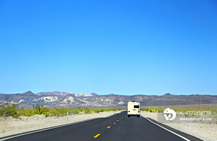 Quiet road - White RV on the empty highway leading to Death Valley in California, surrouned by sandy ground with bushes, mountains in the background.