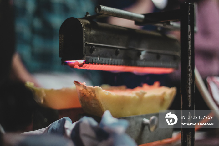 Detail of a raclette heater. Gourmet food being prepared under a gas heater above cheese.