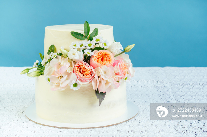 appetizing creamy white two-tiered wedding cake decorated with fresh flowers on a table with a lace tablecloth in studio on a blue background