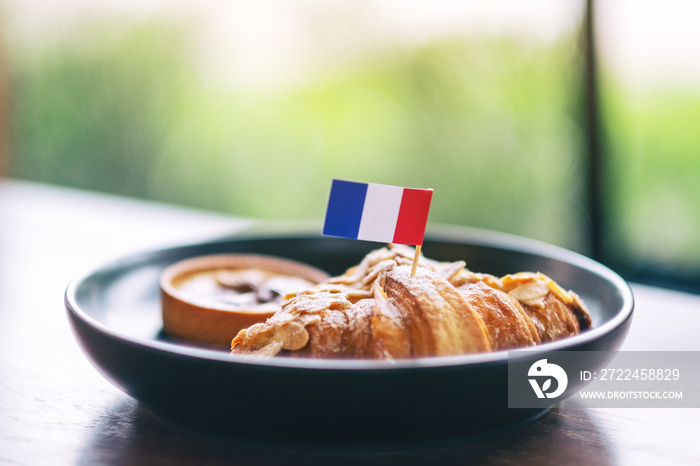 A piece of croissant and tart with a flag of France in a black plate on wooden table in cafe