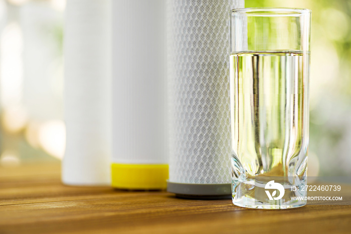 A glass of purified water and filter cartridges on wooden table on green natural background