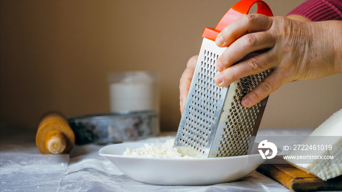 Close-up hands of mature woman is rubs a white soft cheese on a steel grater for preparing bakery at home kitchen