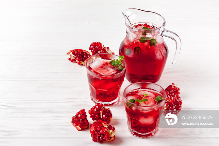 Pomegranate cocktail with ice and mint in beautiful glasses and jug, fresh ripe pomegranate on white wooden background. Sweet red juice. Close up photography. Selective focus