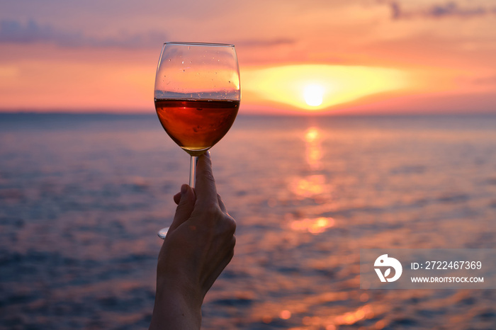 A glass of red wine in the hands of a girl in the rays of the sunset on the seashore. Alcoholic beverage tasting. Beautiful sunset with wine on the beach. Romantic evening aperitif. Glass closeup