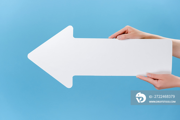 Close up cropped shot of beautiful female hands holding big white paper arrow, isolated on blue studio background wall with copy space for advertisement. Advertising area, mockup