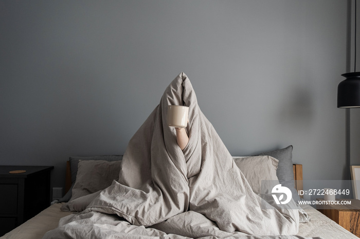 Woman sitting in bed, holding mug with coffee in hand. Happy morning breakfast concept. Freelancer, pandemic quarantine day lifestyle concept.