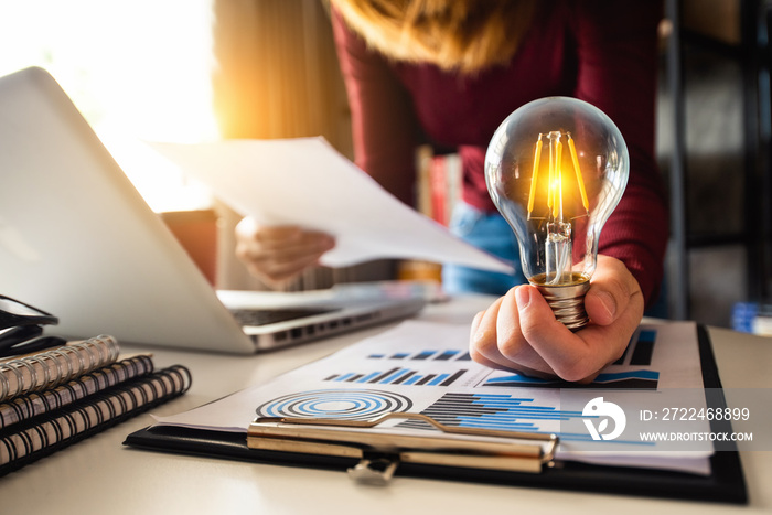 business hand holding lightbulb with using laptop computer and money stack in office. idea saving energy and accounting finance concept  in morning light