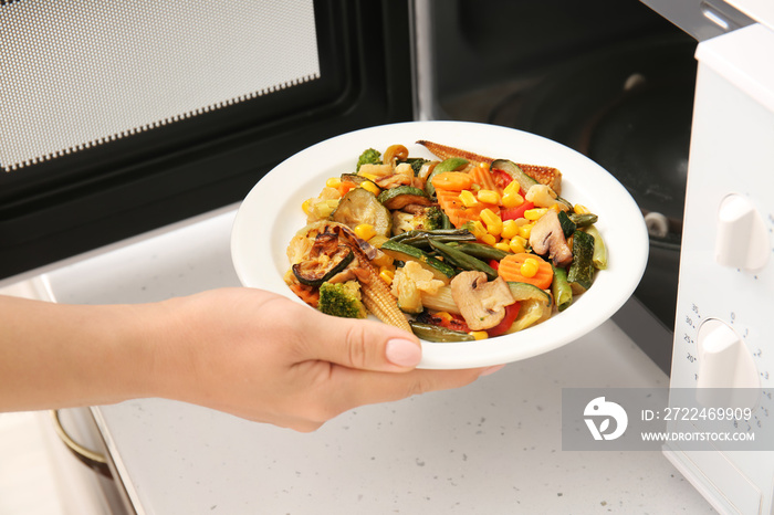 Woman putting plate with vegetables into microwave oven in kitchen, closeup