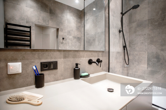 Bathroom in a modern style with gray tiles. Large mirror. Bathtub with shower and glass partition, towel rack and hanger.