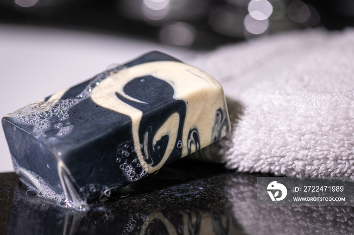 A sudsy bar of black and white soap net to a fluffy towel on a reflective black granite bathroom countertop with the sink out of focus in the background.