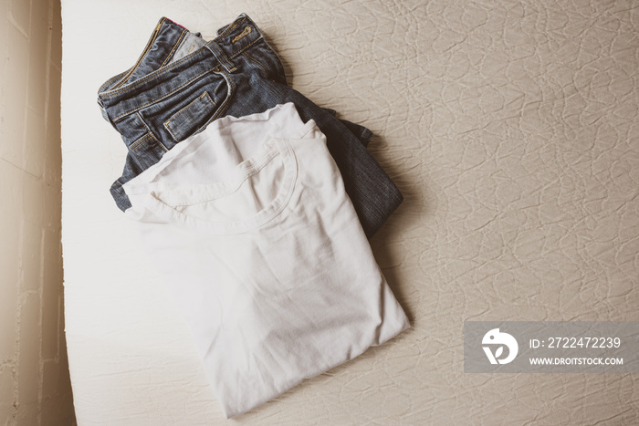 HIgh angle view of folded plain white t-shirt and blue jeans on edge of cream coloured bedding - warm matte filter effect