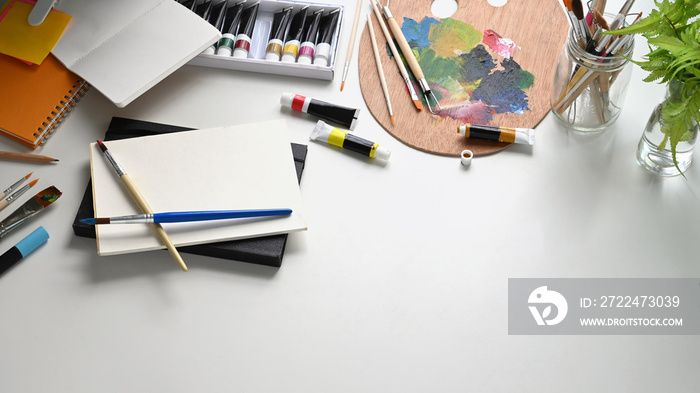 Top view image of artist/painter working table with accessories putting on it. Flat lay stack of notebooks, Color palette, Paint brush vase, Paint brushes, Oil color tube. Orderly workplace.