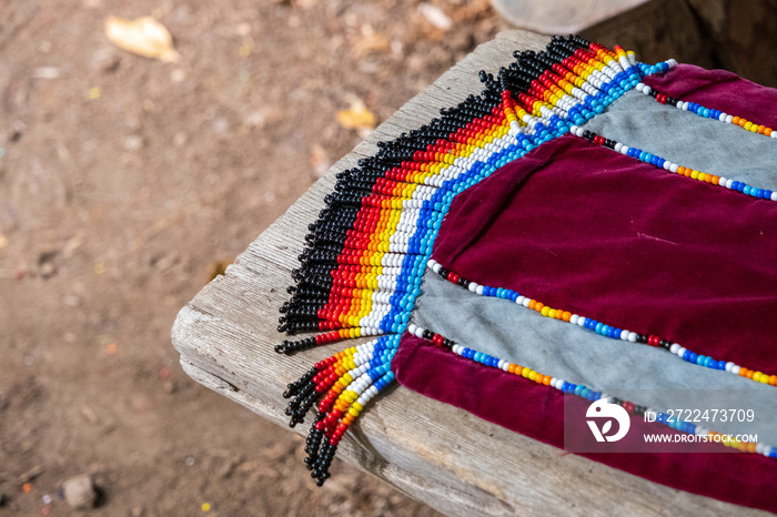 Native American beadwork fringe adorns a crimson and gray scarf outdoors on a wooden bench.