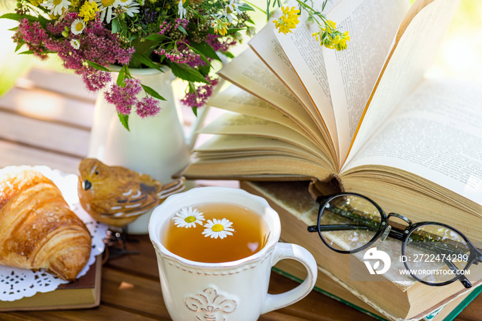 Mug of herbal tea of chamomile, bouquet of meadow flowers, croissant, books on wooden table. Rest in garden, healthy drink herbal tea, reading books, breakfast, vacation, summertime mood concept