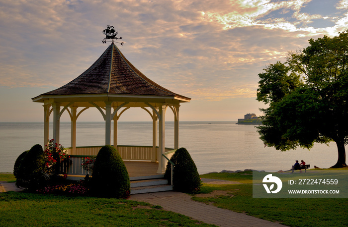 The gazebo at Queens Royal Park, Niagara on the Lake, photographed at sunrise in the summer looking out over Lake Ontario and Fort Niagara with a couple drinking coffee on a bench under a tree