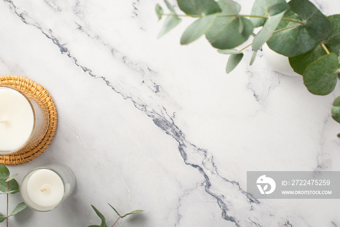 Top view photo of candles on rattan serving mat and ceramic vase with eucalyptus on white marble background with copyspace