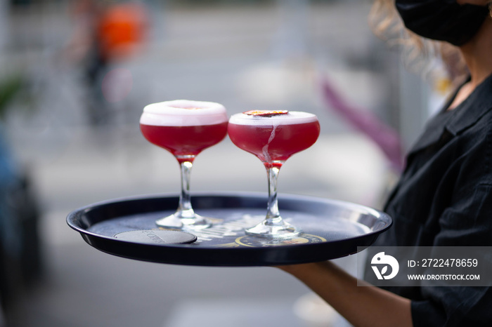 masked waitress carries two cosmopolitan cocktails on a tray