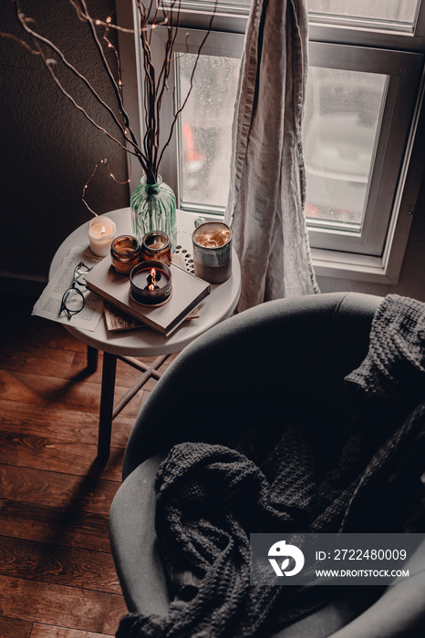 Cozy corner in the living room. Cozy warm blanket. Winter time. Candles on the table. Scandinavian interior decor. Cold winter time. Grey chair, throw blanket. Reading a book. Warm and cozy home