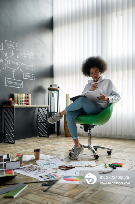 Looking for fresh ideas. Young afro american designer reading magazine while sitting on the chair in the creative office. Designer’s work tools and equipment lying on the floor. Design studio