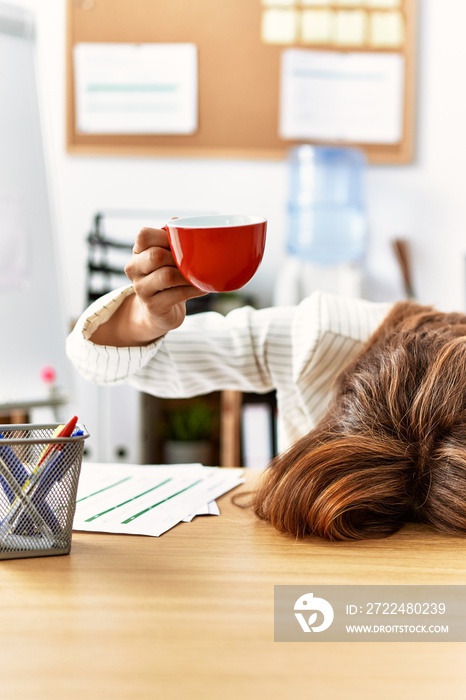 Young hispanic businesswoman overworked with head on table and holding cup of coffee at the office.