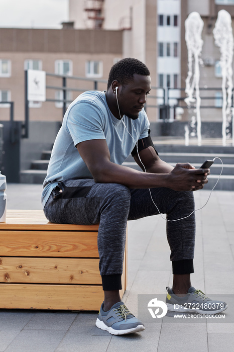 Young muscular Black man in sportswear sitting on bench outdoors, listening to music through earphones after workout