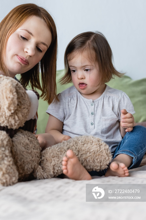 Parent holding teddy bear near kid with down syndrome on bed.