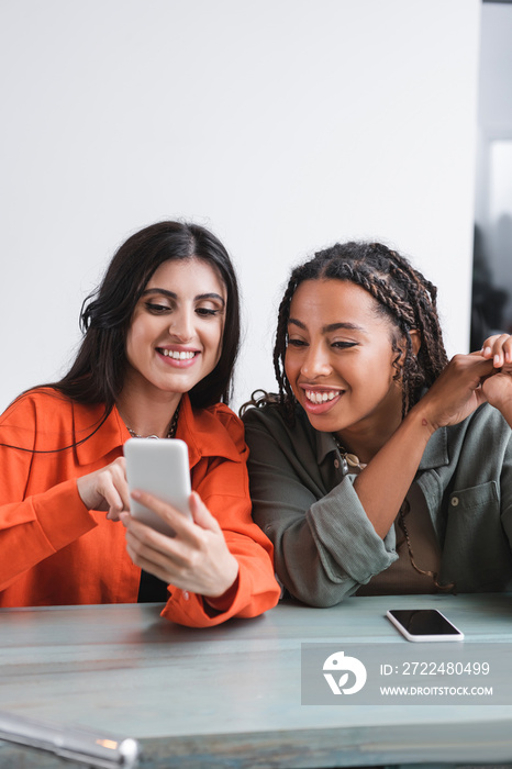 Cheerful african american woman looking at cellphone near friend in cafe