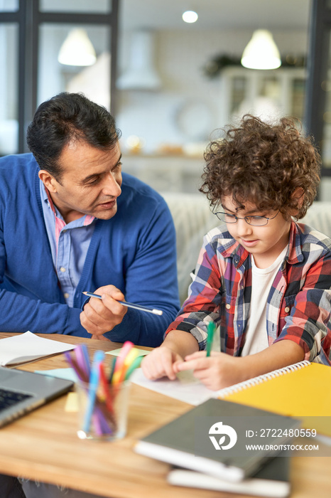 Best teacher. Loving hispanic father helping his son with homework, explaining task while sitting together at the desk at home