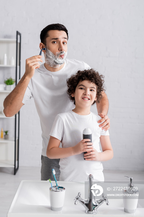 Smiling arabian boy holding shaving foam near father shaving beard in bathroom