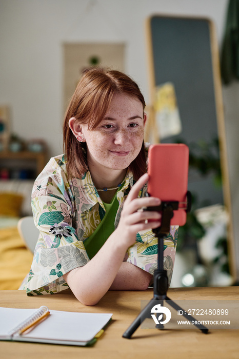 Teenage girl using her smartphone on tripod to recording blog at table in her room