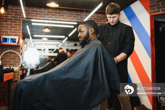 Portrait of young black man being trimmed with professional electric clipper machine in barbershop.Male beauty treatment concept. Young African guy getting new haircut in barber salon