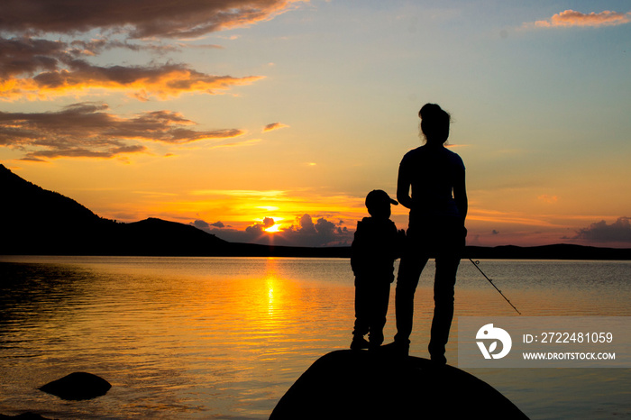 Mom and her little daughter are fishing with a fishing rod on a pier at sunset by the lake