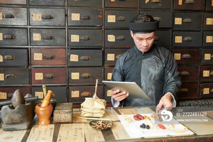 Traditional Asian medicine practitioner reading recipe on tablet computer when making remedy for patient