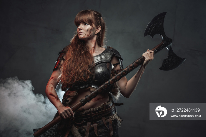 Beautiful and at the same time barbaric female warrior from nord poses in dark foggy background holding two handed axe.