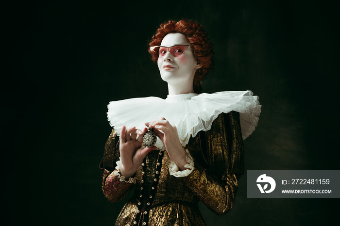 Secrets. Medieval redhead young woman in golden vintage clothing as a duchess in red sunglasses holding a parfume on dark green background. Concept of comparison of eras, modernity and renaissance.