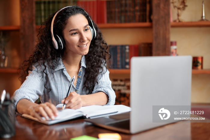 Woman sitting at desk, using laptop and writing in notebook