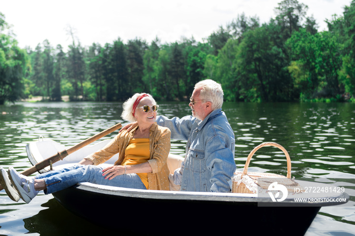 Talk in boat. Calm positive senior woman turning to the husband while sitting in the boat near him