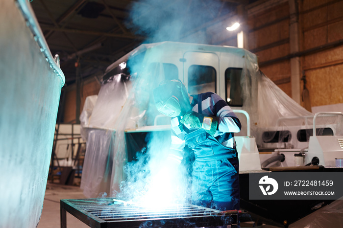 Shipbuilding factory worker in protective mask welding metallic details of one of boats