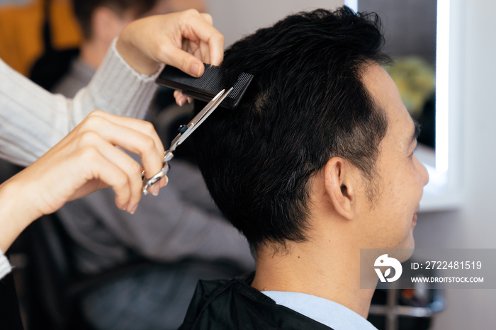 Closeup asian female barber making haircut for male customer with male hairstyle in qualified barbershop. Men’s hairstyling by a scissor and comb in hair salon concept.