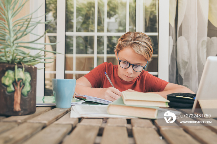 Young student doing homework at home sitting at table outdoor. Boy writing and studying with school books and digital tablet pad. Kid doing a research online. Education technology lifestyle concept.