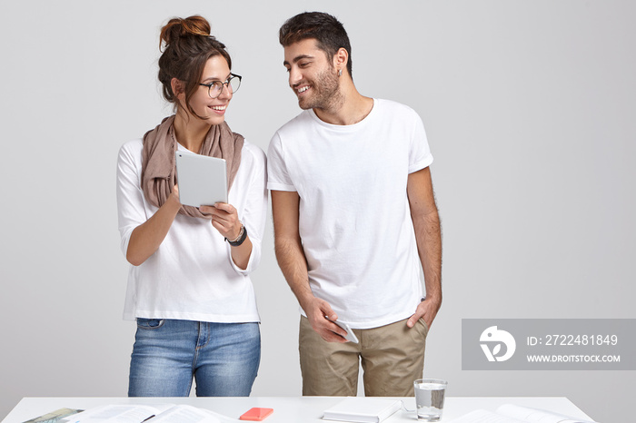 Purposeful business people work on digital tablet, enjoy teamwork, being professional bankers, look happily, stand against white background, pose at table, drink water, dressed in stylish clothes