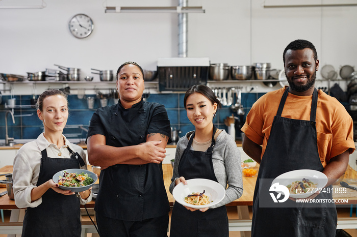 Diverse group of people wearing aprons and holding gourmet dishes in professional kitchen