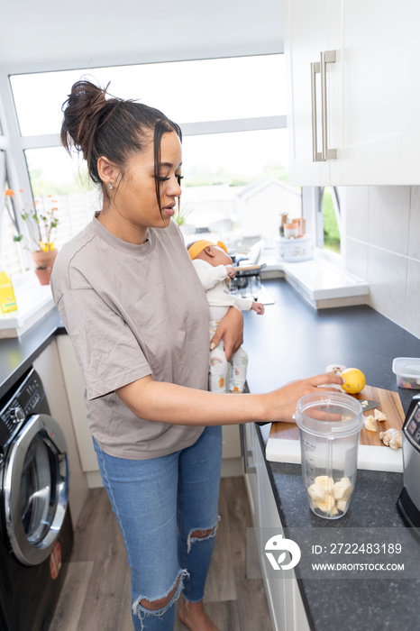 Mother preparing smoothie while carrying baby girl in domestic kitchen