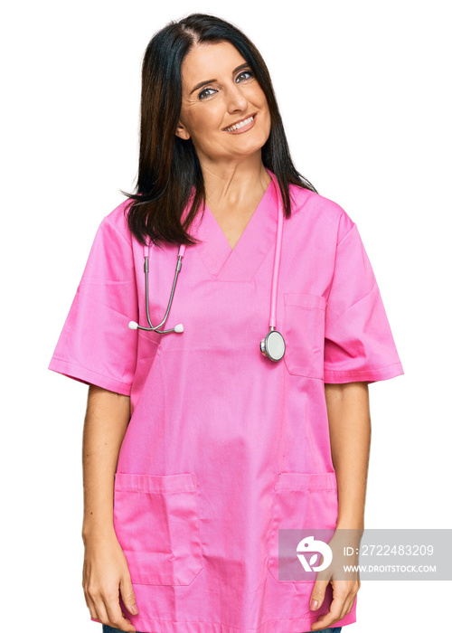 Middle age brunette woman wearing doctor uniform and stethoscope looking positive and happy standing and smiling with a confident smile showing teeth